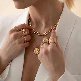 18k gold plated brass Gemini star sign Zodiac Necklace with cubic zirconia stones and intricate detailing on a coin disc pendant and on fine ball chain worn on a model with a gold chunky link chain and multi stacking gold rings, wearing a white Zara blazer.