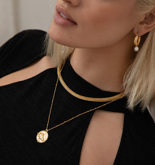 18k gold plated brass Taurus Zodiac Necklace with cubic zirconia stones and intricate detailing on a coin disc pendant and on fine ball chain - worn on a model wearing a black Zara dress and layering the jewellery with gold chubby hoops with a freshwater pearl charm and another gold link cain necklace.