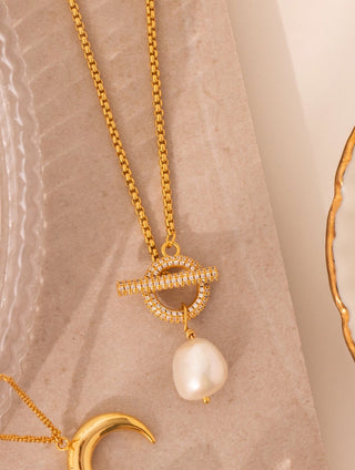 gold plated Cubic Zirconia encrusted T Bar & freshwater Pearl Necklace - Gold Lunar displayed on a concrete tile next to a crescent moon shaped pendant necklace