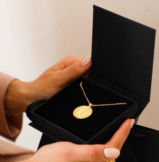 18k gold plated coin pendant necklace with cubic zirconia gemstones around the outside on a fine gold chain displayed inside a gold lunar gift box which is black with a velvet insert. Box is being opened by a model with pale pink gel nails.