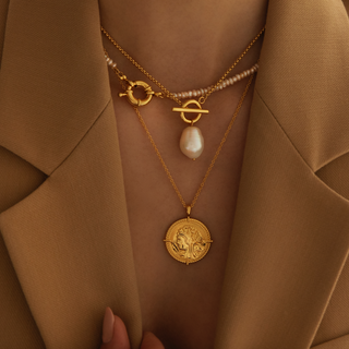 Gold plated ,layering necklace set featuring all freshwater pearl necklace with round clasp, then a t bar and freshwater pearl charm necklace and then a large coin necklace. worn by a model wearing a beige Zara blazer open at the front