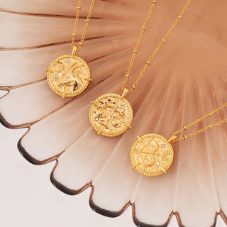 18k gold plated brass Libra star sign Zodiac Necklace with cubic zirconia stones and intricate detailing on a coin disc pendant and on fine ball chain - displayed on a pink flower plate with the Capricorn and Sagittarius necklaces 