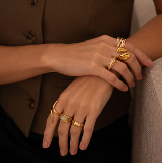 model wearing a brown Zara waistcoat resting an arm on the side of a sofa. wearing multiple rings across fingers including a wrap cross over ring, chunky wrap ring, cubic zirconia encrusted ring, gemstone jade ring, band ring.